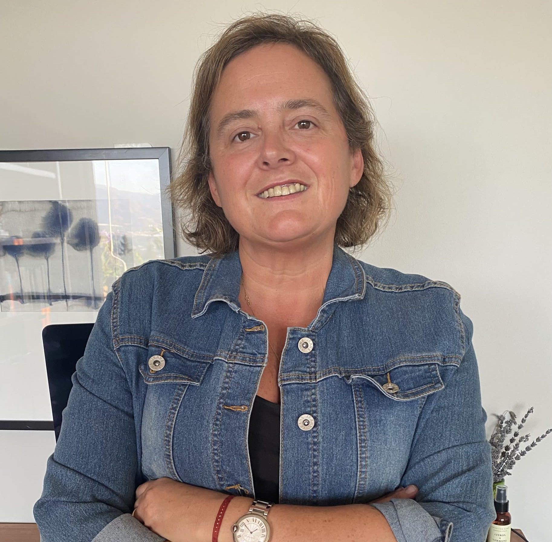 Smiling woman in a denim jacket with arms crossed, standing at a desk in a bright office. (Informative portrait)