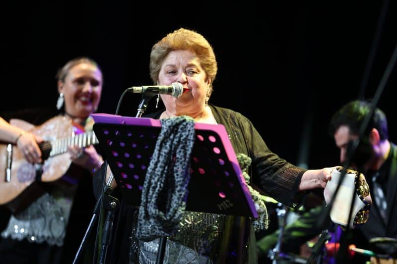 Older woman singer at a microphone on stage with a guitarist in the background, music stand with purple sheet music in front.