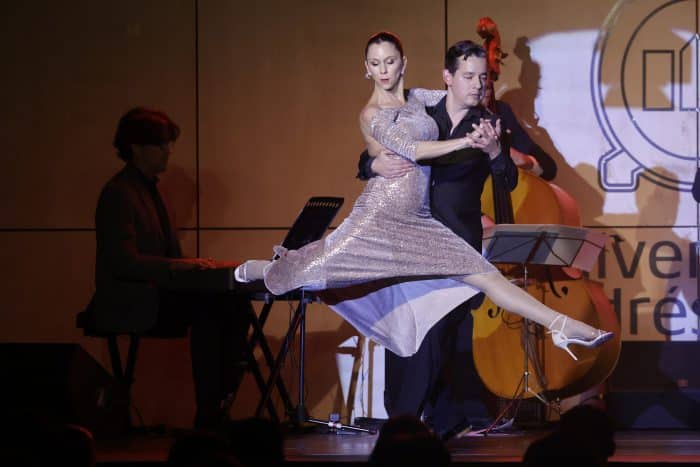Couple dancing a dramatic lift on stage: woman in a sparkling silver dress leaps mid-air while her partner supports her, with a pianist in the background.