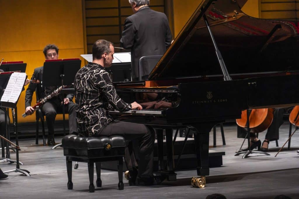 Pianist in a patterned shirt playing a Steinway grand piano on a concert stage, with a conductor and seated musicians in the background.