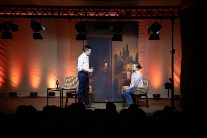Two actors on a stage set as a living room—man standing with a glass facing a seated woman; warm orange lighting and furniture.