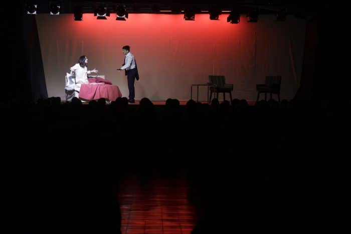 A stage scene with a seated person at a pink‑tablecloth table facing a standing man reaching toward them, under red stage lights. A few chairs sit to the right; audience silhouettes are in front.