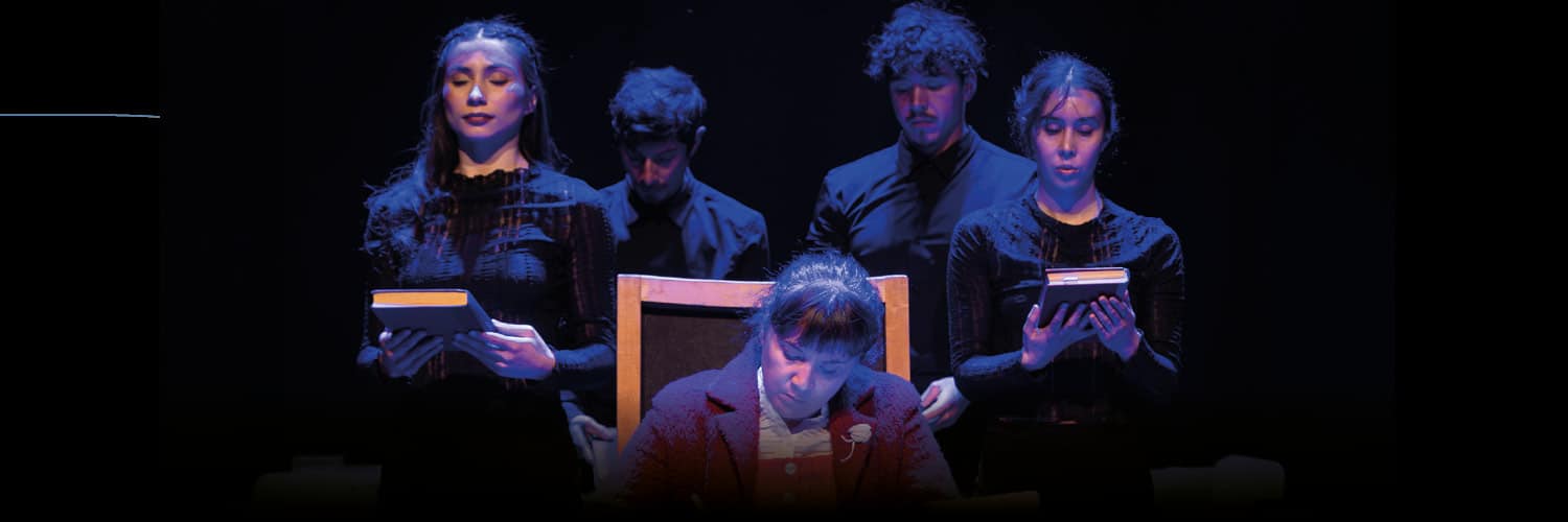 Theatrical stage scene: a woman in a red jacket sits at a chair while four performers in black stand behind her, each holding a book under blue stage lighting.