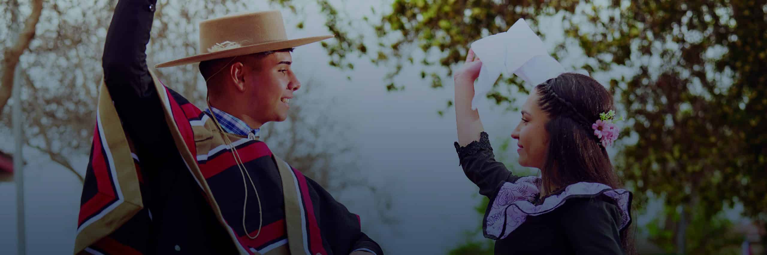 Two people in traditional attire dance outdoors, the man in a wide-brim hat and poncho facing a woman who raises a paper hat.