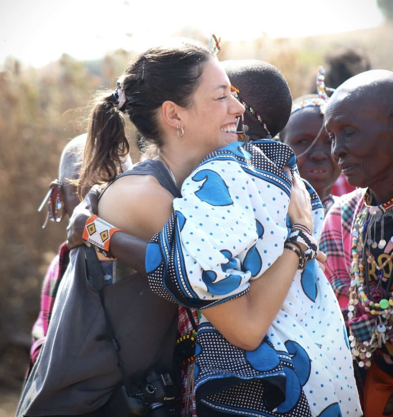 Woman smiling while hugging a baby wrapped in a blue and white patterned blanket; several people in colorful beaded clothing nearby outdoors.