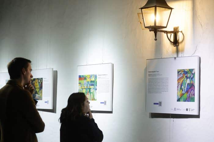 Two people stand in a dim gallery, studying framed colorful artworks hung on a white wall beside a warm wall lantern.