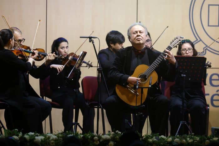 Man with guitar performing on stage surrounded by an orchestra (violinists and musicians) and sheet music stands.