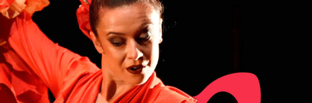 Female flamenco dancer in a red dress performs on stage, arm raised against a dark background.