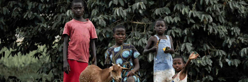 Four children stand outdoors in front of dense foliage, with a light-brown goat at their feet near the center.