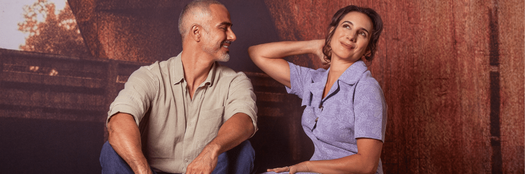Man and woman sit side by side, smiling at each other against a rust-colored wall.