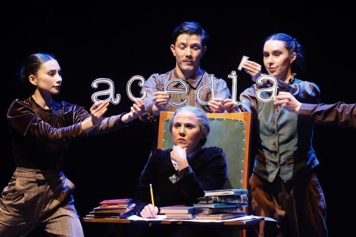 A theater scene: an older woman sits at a desk with books, writing while others hold letters spelling a word above her head.