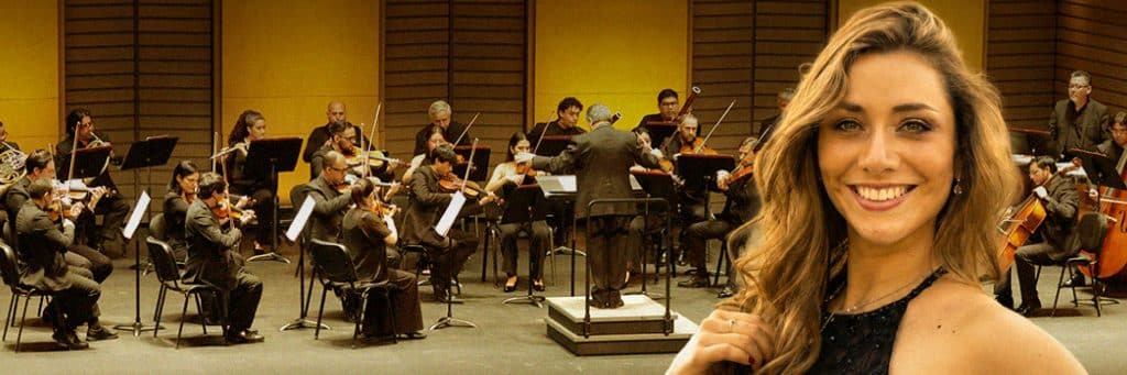 Orchestra performing on stage with a smiling woman in the foreground (right).