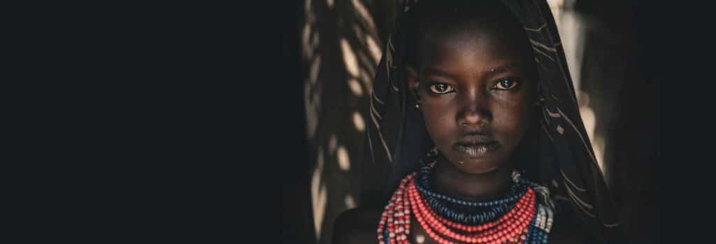 Close-up portrait of a young African girl wearing layered red, blue, and white bead necklaces, gazing directly at the camera with a serious expression.]