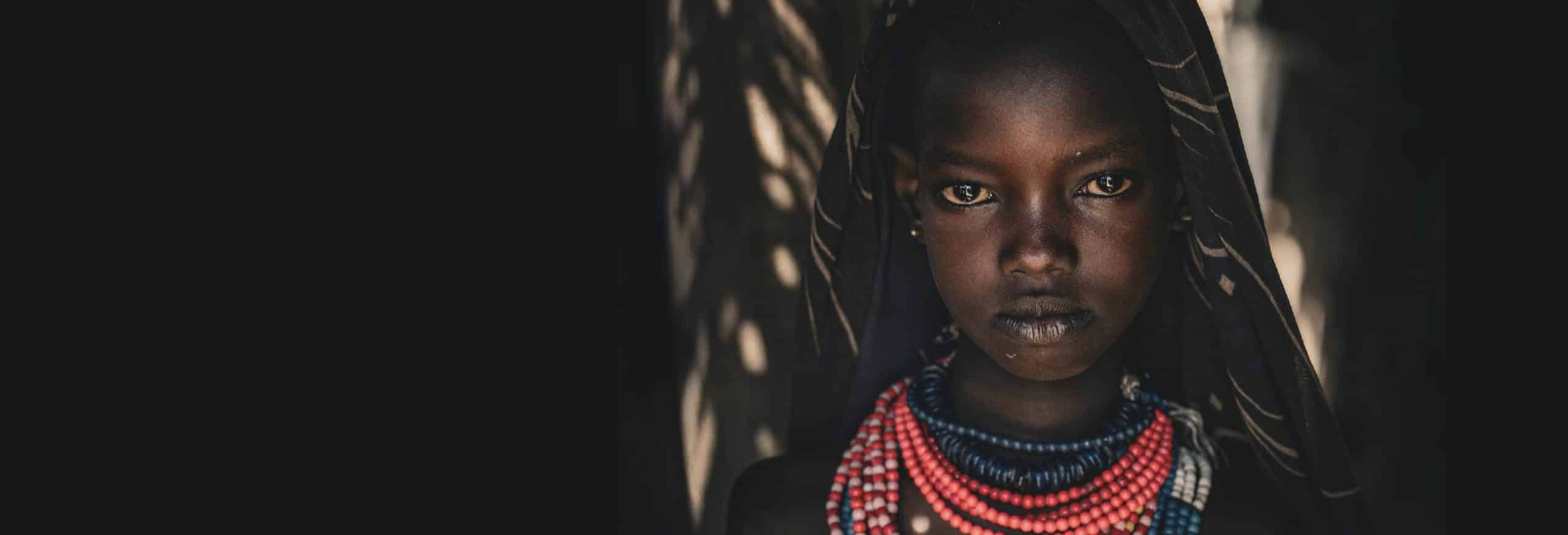 Close-up portrait of a young African girl wearing layered red, blue, and white bead necklaces, gazing directly at the camera with a serious expression.]