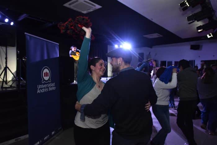 Couple dancing together at a campus celebration, with a blue Universidad Andrés Bello banner and a guitarist performing on stage nearby.